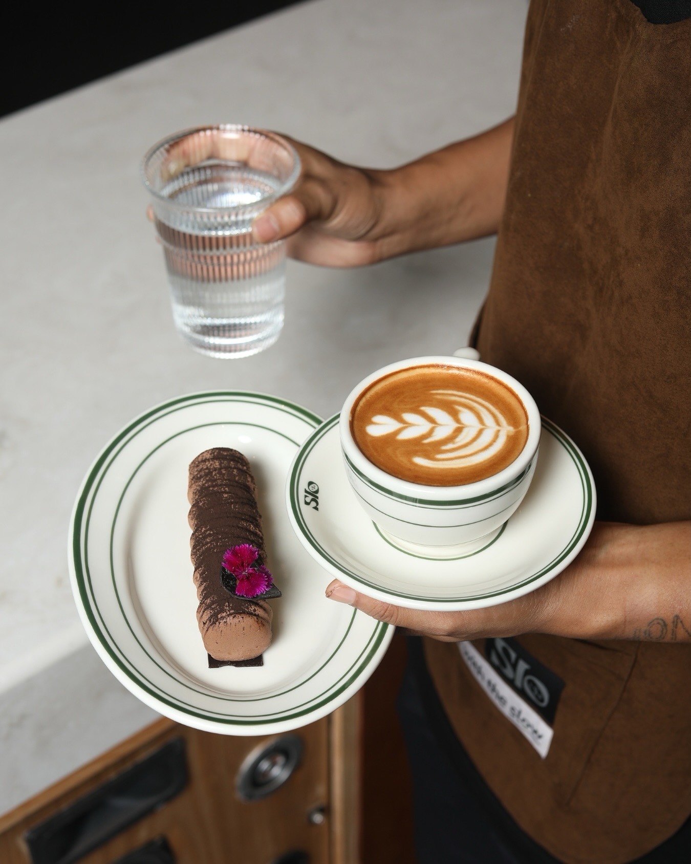 Flat white with rosetta latte art on a green-rim Slo cup, chocolate dessert alongside